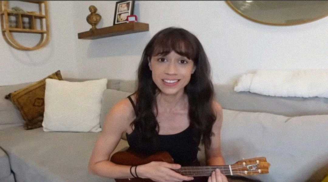 A woman with long brown hair sits on a light-colored couch, smiling and holding a small ukulele. There are pillows behind her and shelves with decorations on the wall.