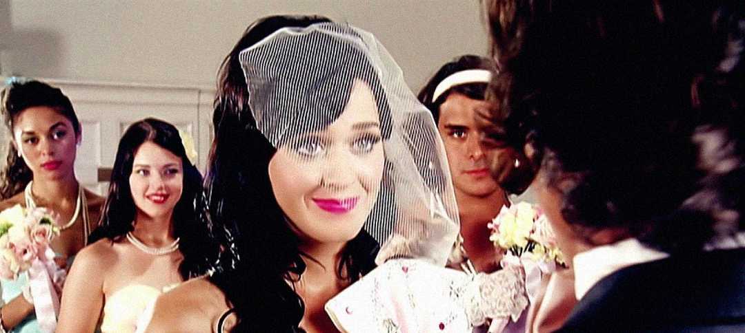A bride with dark hair and a veil smiles while facing a groom. Three bridesmaids holding bouquets stand behind her, dressed in pastel colors, with one looking directly at the camera.