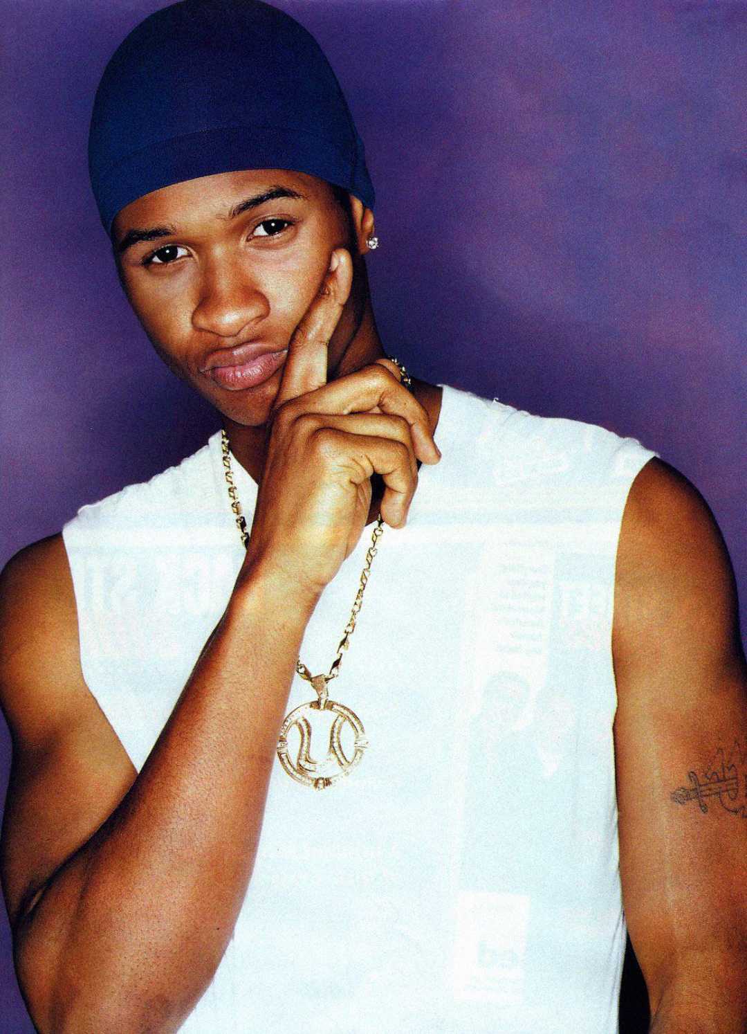 A young man wearing a blue durag, white sleeveless shirt, and gold chain poses against a purple background, resting his chin on his hand and slightly pouting.