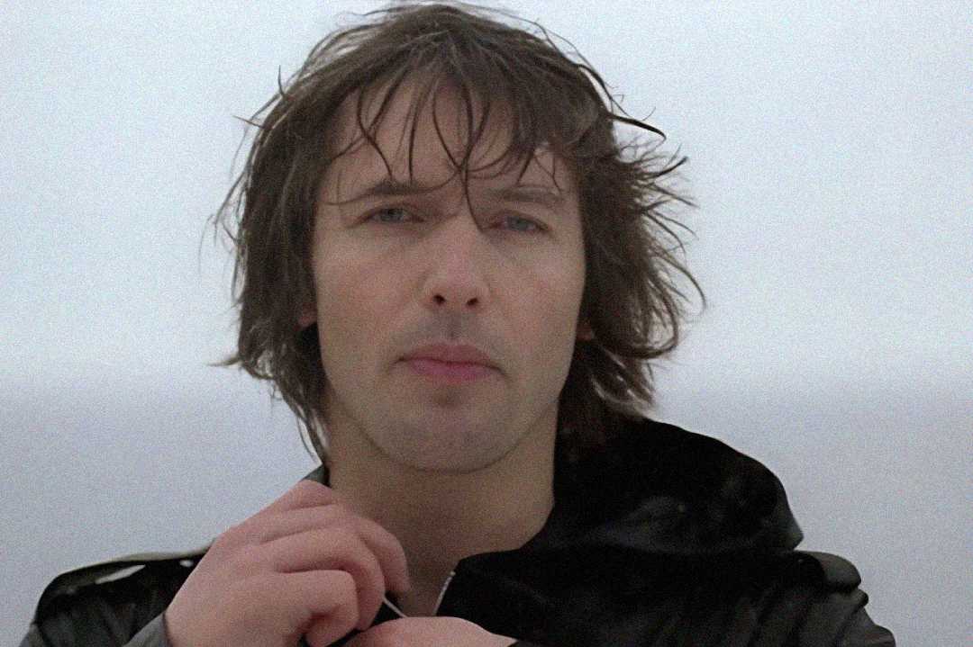 A person with wet, tousled hair stands outdoors in front of a foggy, gray background, wearing a dark jacket and looking directly at the camera while adjusting the collar.