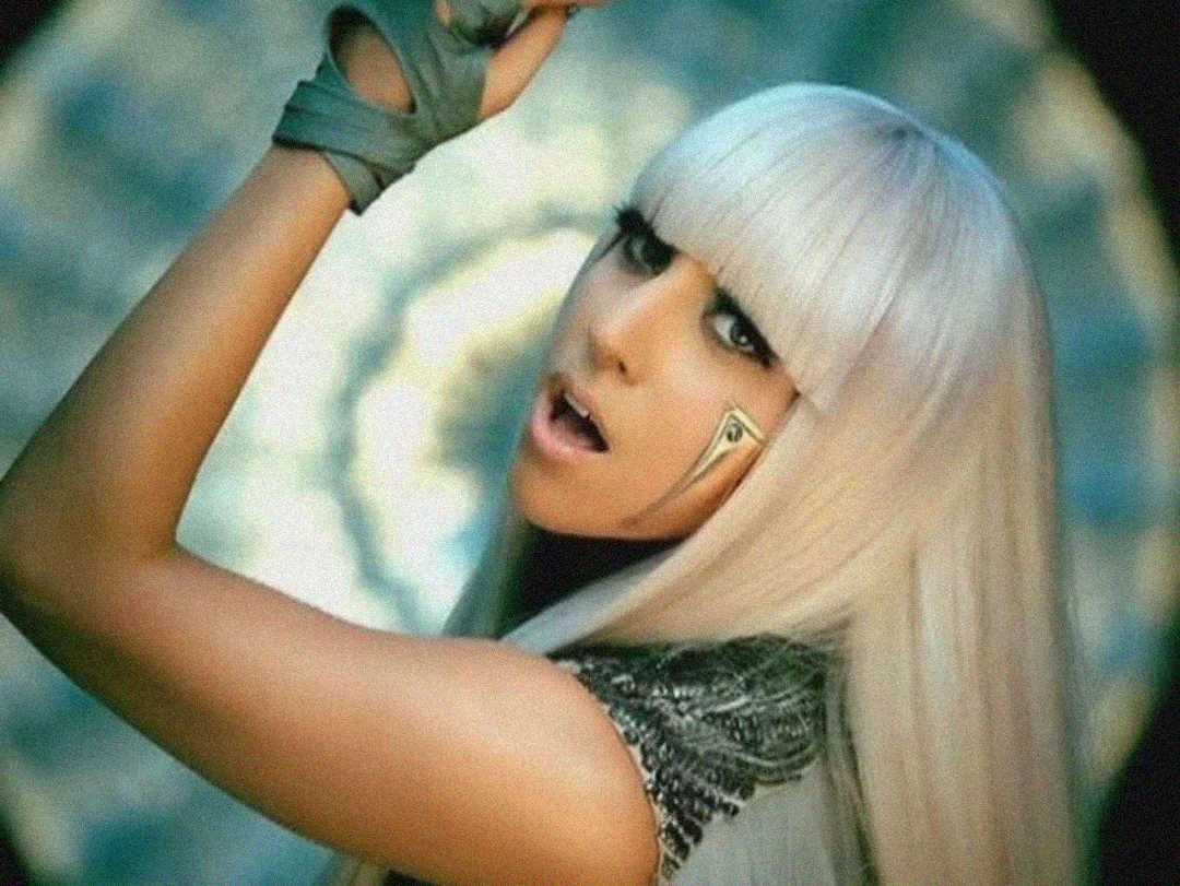 A person with long platinum blonde hair and blunt bangs poses with their arm raised. They have dramatic eye makeup and a metallic accent on their cheek, wearing a futuristic, silver-toned outfit against a circular, glowing background.
