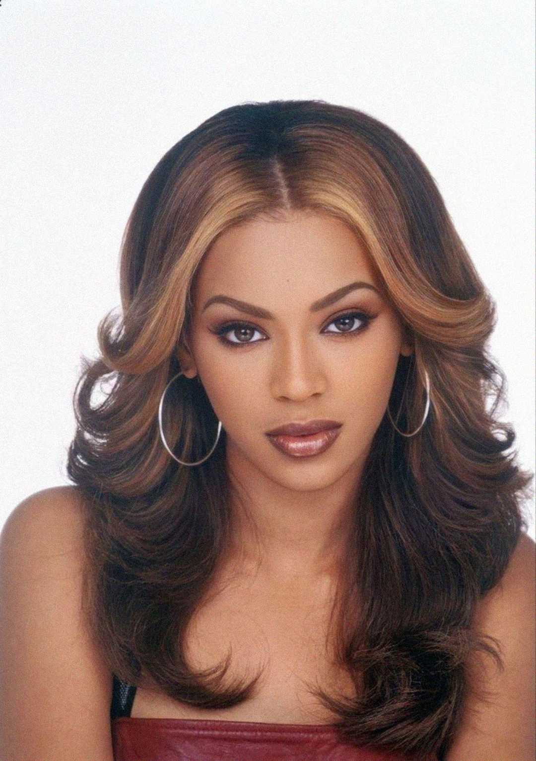 A woman with long, wavy, light brown hair with blonde highlights, wearing hoop earrings and a maroon top, looks directly at the camera with a neutral expression against a plain background.