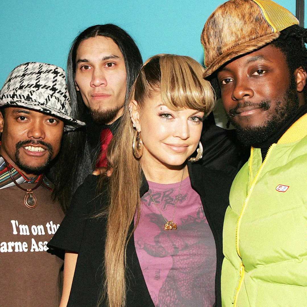 Four people pose together and smile at the camera. One woman stands between three men. They wear colorful and patterned clothing and hats, standing against a plain light blue background.