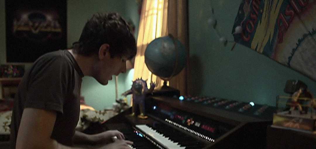 A young person plays an electronic keyboard in a dimly lit room decorated with posters, a globe, and figurines on the desk. Light from a window shines behind them.