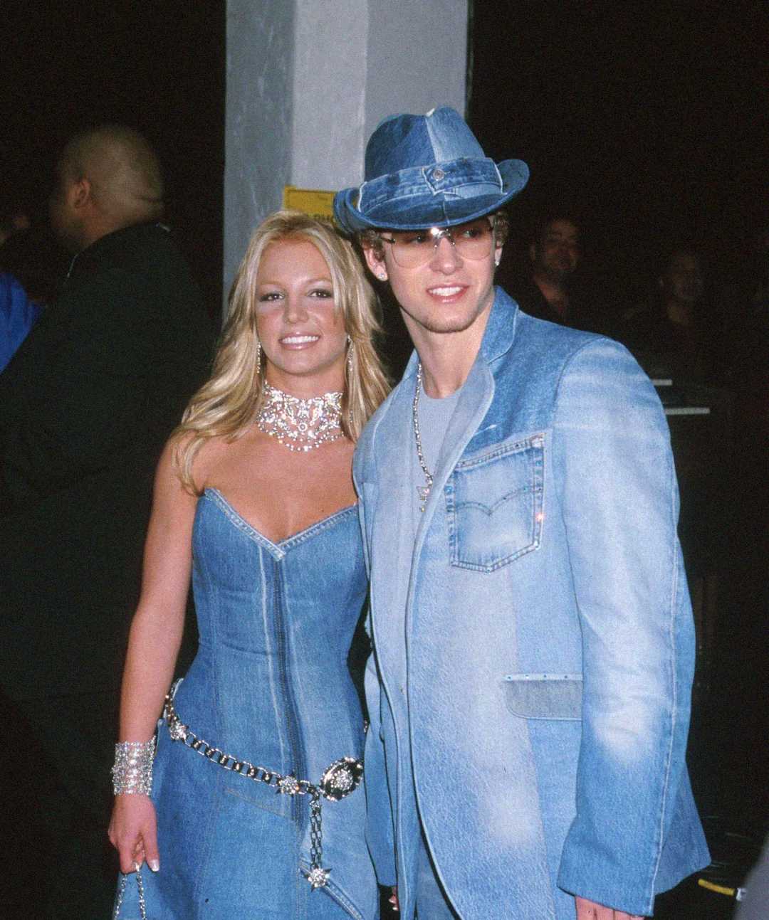 A woman and a man pose together at an event, both dressed in matching blue denim outfits. The woman wears a strapless denim dress with silver jewelry, while the man wears a denim suit, hat, sunglasses, and a white shirt.