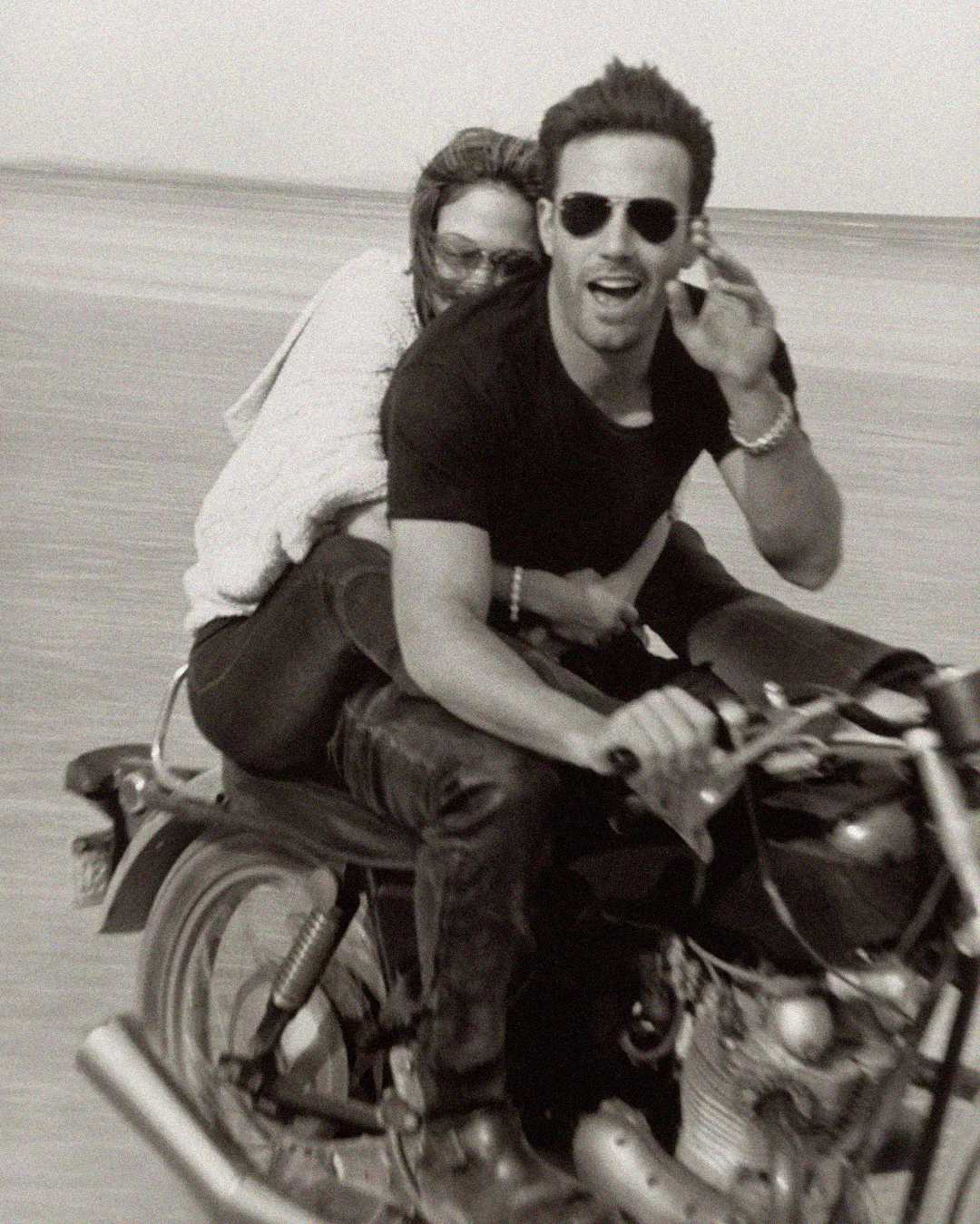 Black and white photo of a smiling man in sunglasses and a black t-shirt riding a motorcycle with a woman holding onto him from behind. They are on a sandy beach.