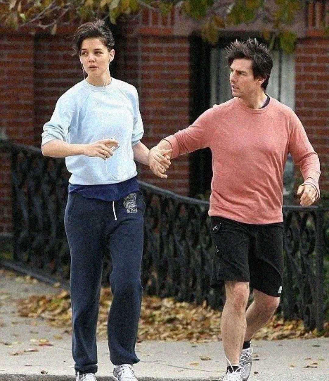 Two people walk outdoors on a sidewalk. The person on the left wears a light blue sweatshirt and dark sweatpants, holding earbuds. The person on the right wears a pink long-sleeve shirt and black shorts. They are holding hands.