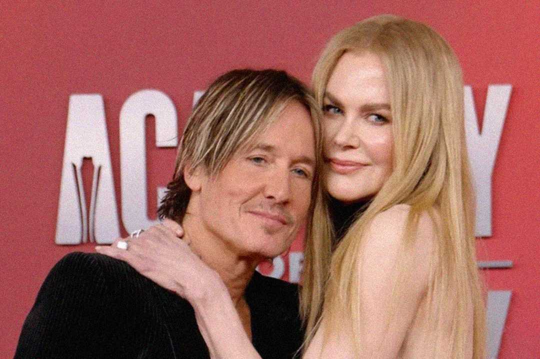 A man and a woman with long blond hair pose together, smiling, in front of a red backdrop with white text. The woman has her arm around the man's shoulder.