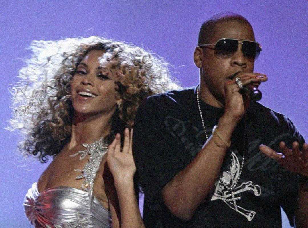 A woman in a silver strapless dress with curly hair and a smiling expression stands beside a man in sunglasses and a black T-shirt as he sings into a microphone against a purple background.