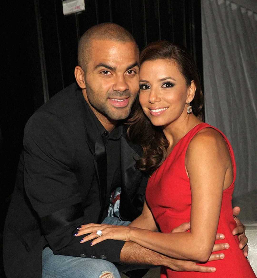 A man in a black jacket and a woman in a sleeveless red dress smile and pose together, seated closely with their arms around each other. The background is dimly lit.