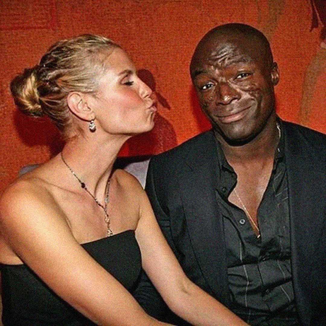 A woman with blonde hair in an updo leans in to kiss a smiling man in a dark suit. They are seated closely together against a warm, reddish background.