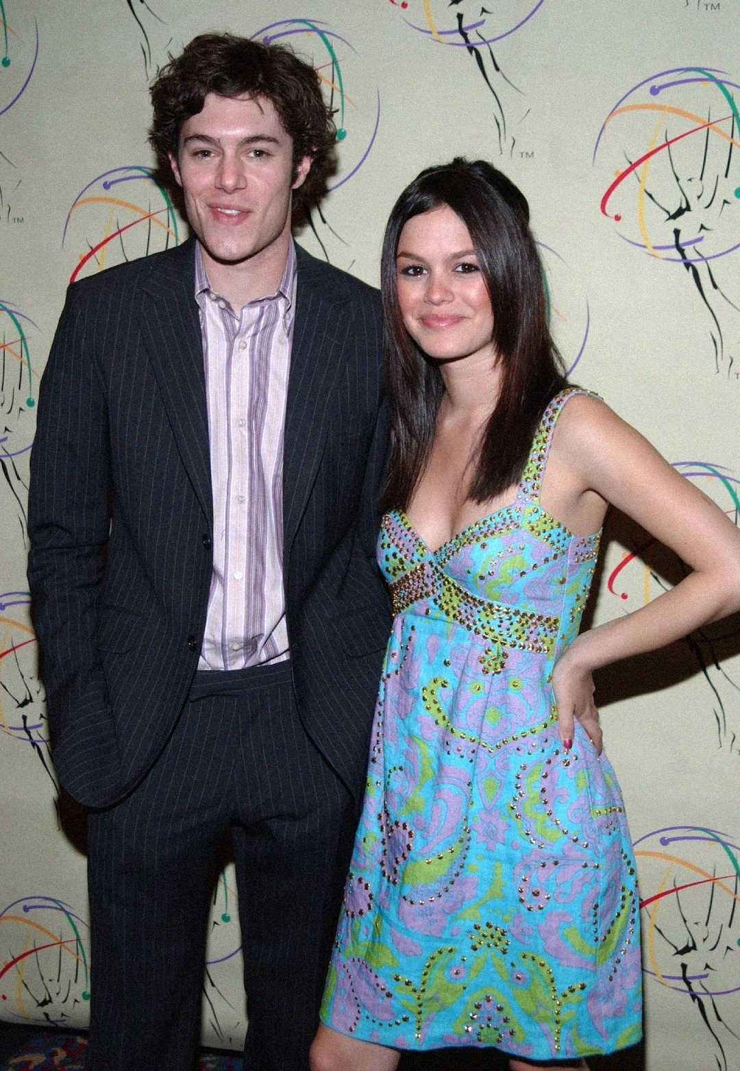 A young man in a pinstripe suit and a young woman in a colorful patterned dress pose together, smiling in front of a cream backdrop with abstract designs.