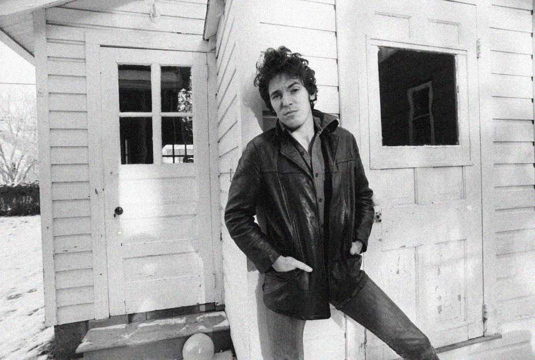 A person with curly hair wearing a leather jacket stands with hands in pockets, leaning against the wall of a white wooden house on a snowy day. Another building with windows and a door is in the background.