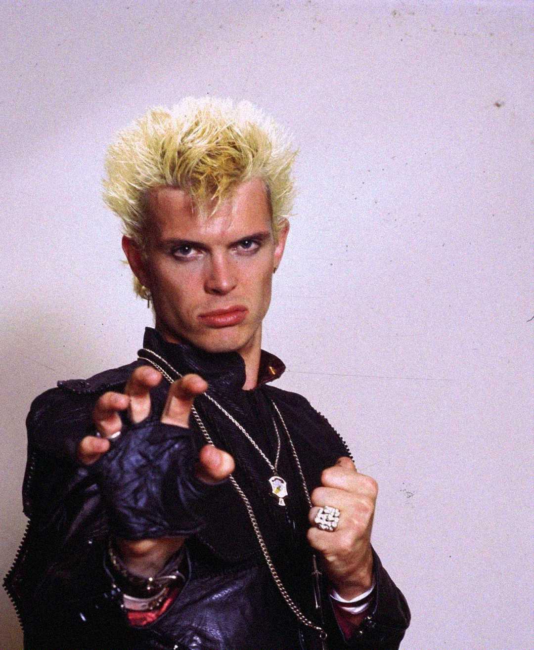 A man with spiky, bleached blonde hair and a black leather jacket strikes a fierce pose, extending one gloved hand forward and clenching the other into a fist, standing against a plain light background.