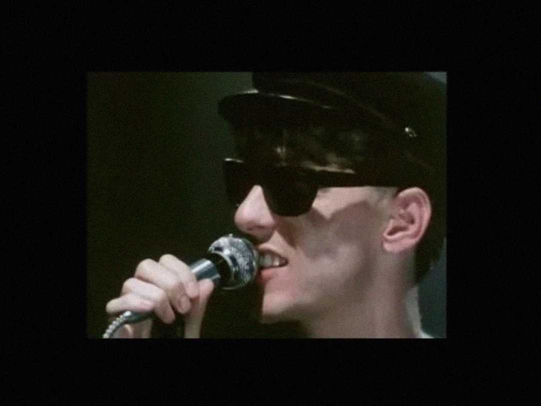 A person wearing dark sunglasses and a black hat sings into a microphone, set against a dark background.