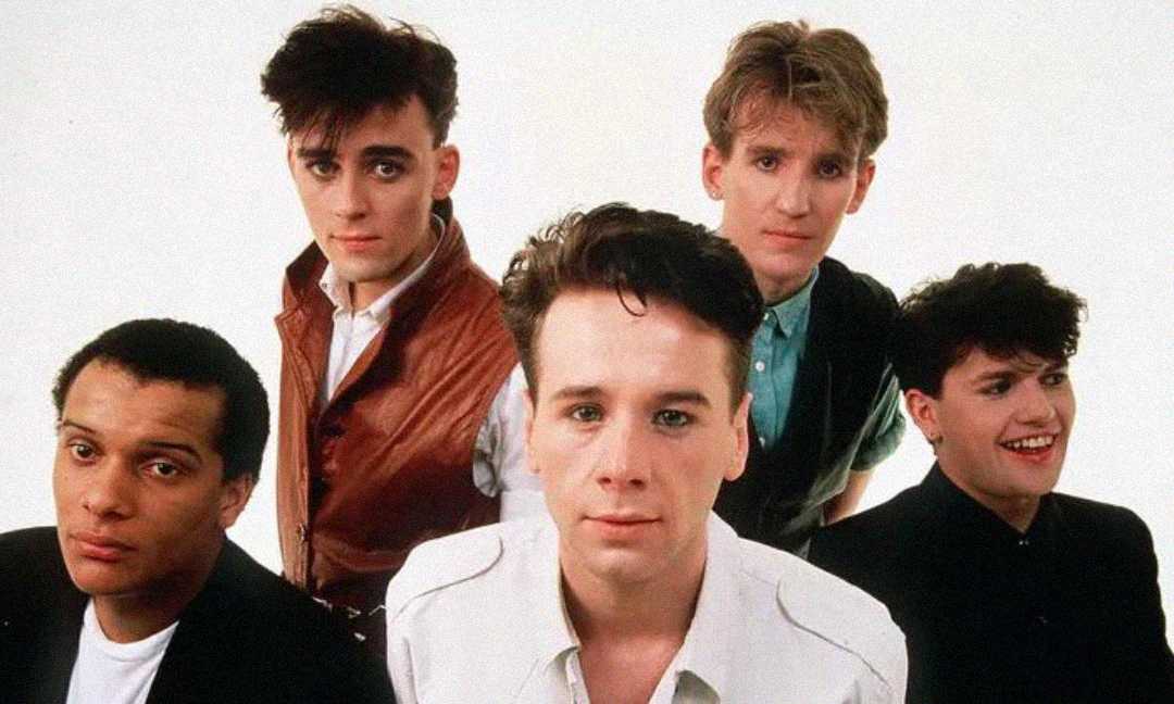 Five men pose together against a plain white background, all with short 1980s hairstyles and casual clothing, looking directly at the camera with neutral or slight smiles.