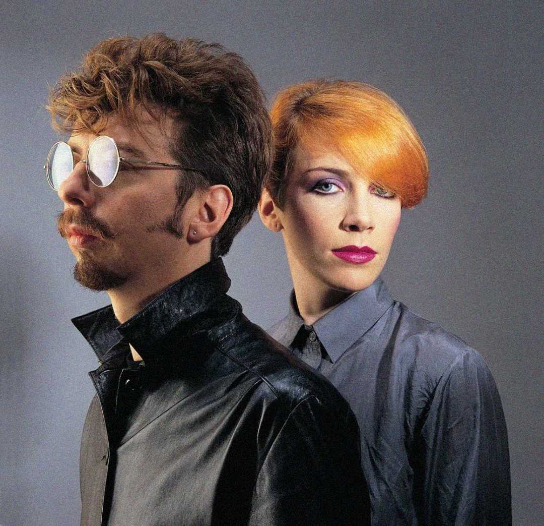 A man with curly hair, a goatee, and round glasses stands next to a woman with orange hair, colorful makeup, and a serious expression. Both wear dark, collared shirts against a plain gray background.