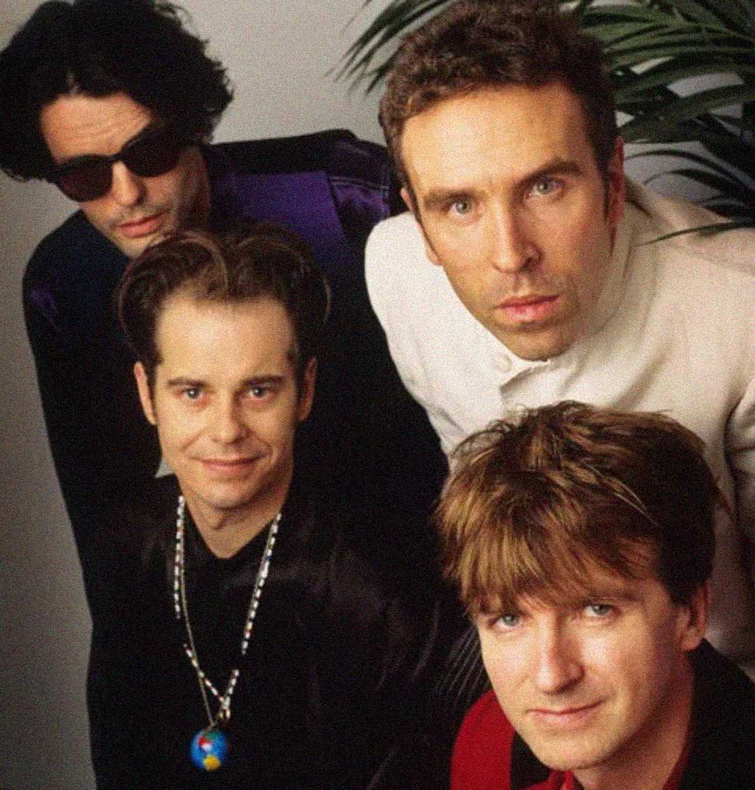 Four men pose closely together, two standing at the back and two seated in front. The man on the left wears dark sunglasses, while the other three look directly at the camera, all with relaxed expressions. A plant is in the background.