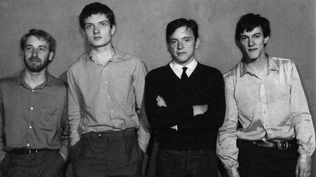 Four young men stand in a row against a plain wall, dressed in collared shirts and trousers, with neutral expressions; the photo is black and white.