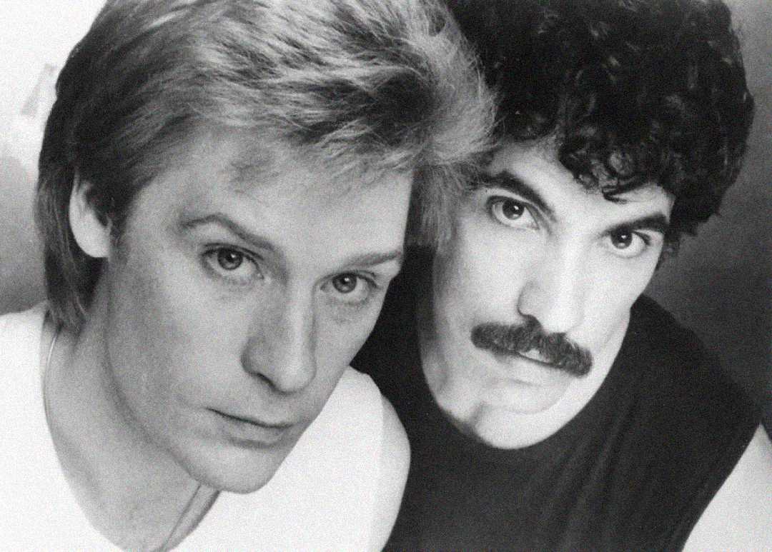 Two men with serious expressions pose closely together. One has light hair and fair skin, while the other has curly dark hair, a mustache, and darker eyes. The image is in black and white.