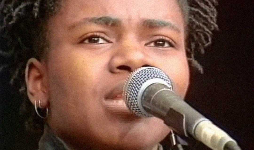 A person with short, twisted hair and an earring is singing or speaking into a microphone, shown in a close-up shot with a dark background.