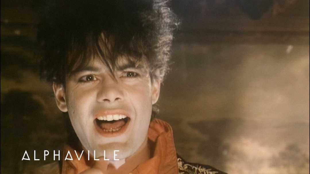 A person with dramatic makeup and wild dark hair sings passionately. The word "ALPHAVILLE" appears in white text in the lower left corner. The background is dimly lit and smoky.