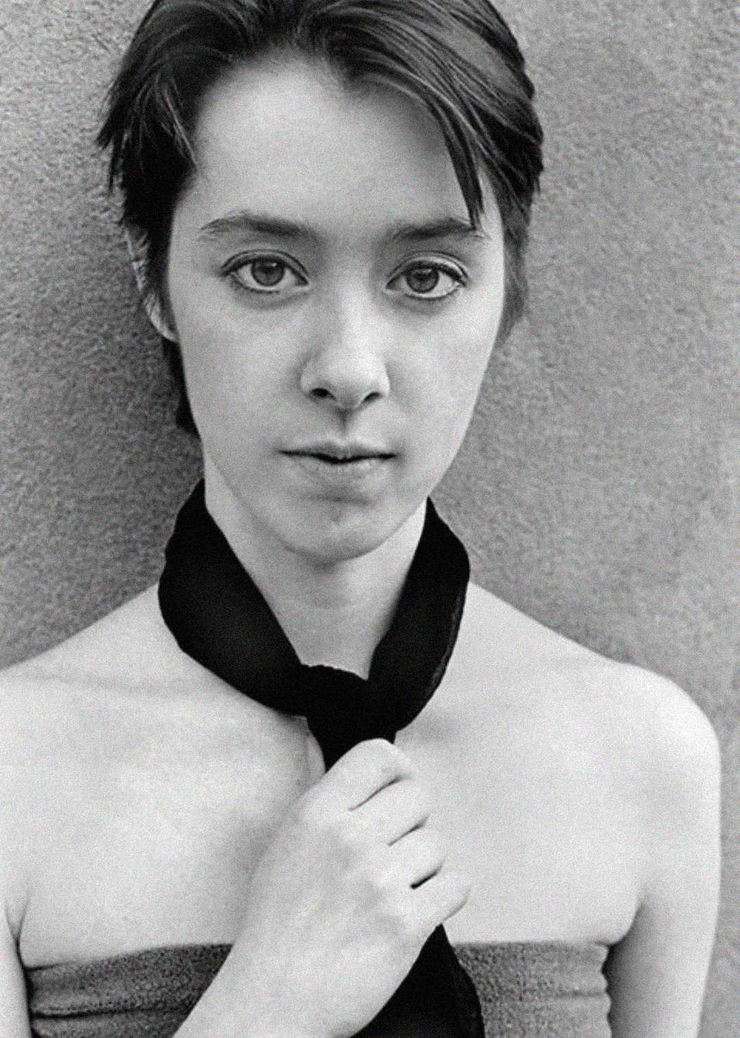 Black and white portrait of a young person with short hair, looking directly at the camera. They wear a strapless top and loosely hold a dark necktie in front of their chest against a plain background.