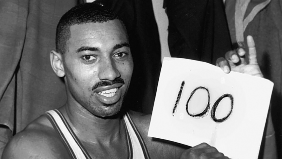 A basketball player sits in a locker room, smiling and holding a piece of paper with &ldquo;100&rdquo; written on it, celebrating a significant achievement. Wilt Chamberlain.