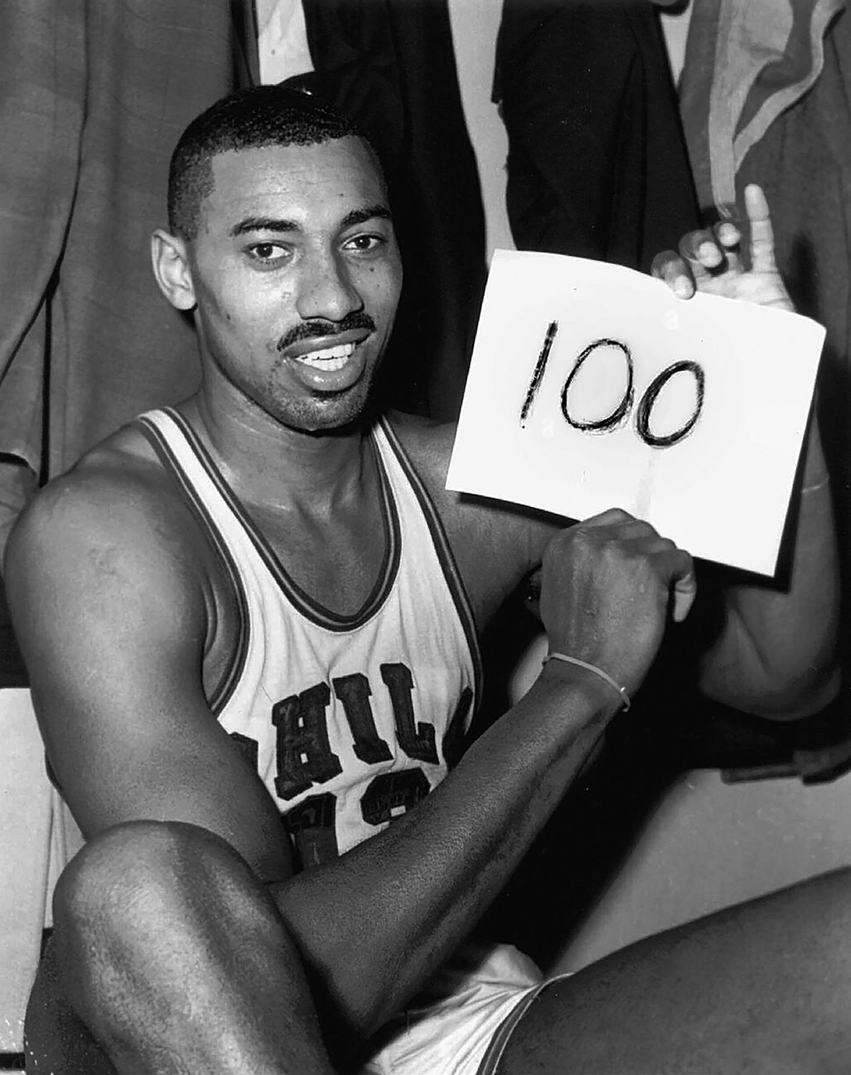 Record. A basketball player sits in a locker room, smiling and holding a piece of paper with “100” written on it, celebrating a significant achievement. 
1. Wilt Chamberlain’s 100-Point NBA Game