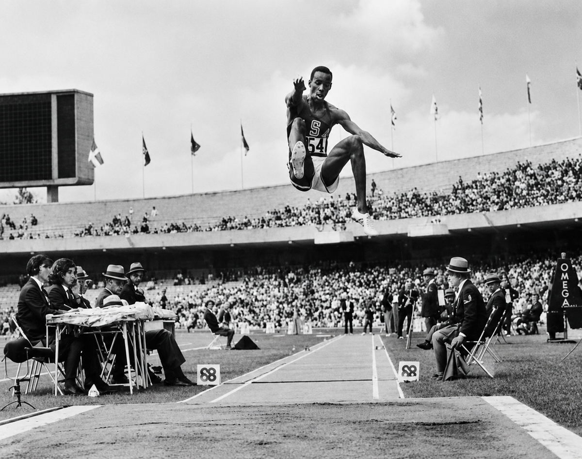 A male athlete in mid-air during a long jump at a stadium event, with spectators in the stands and officials seated on both sides of the runway. 10. Bob Beamon’s 8.90-Meter Olympic Long Jump