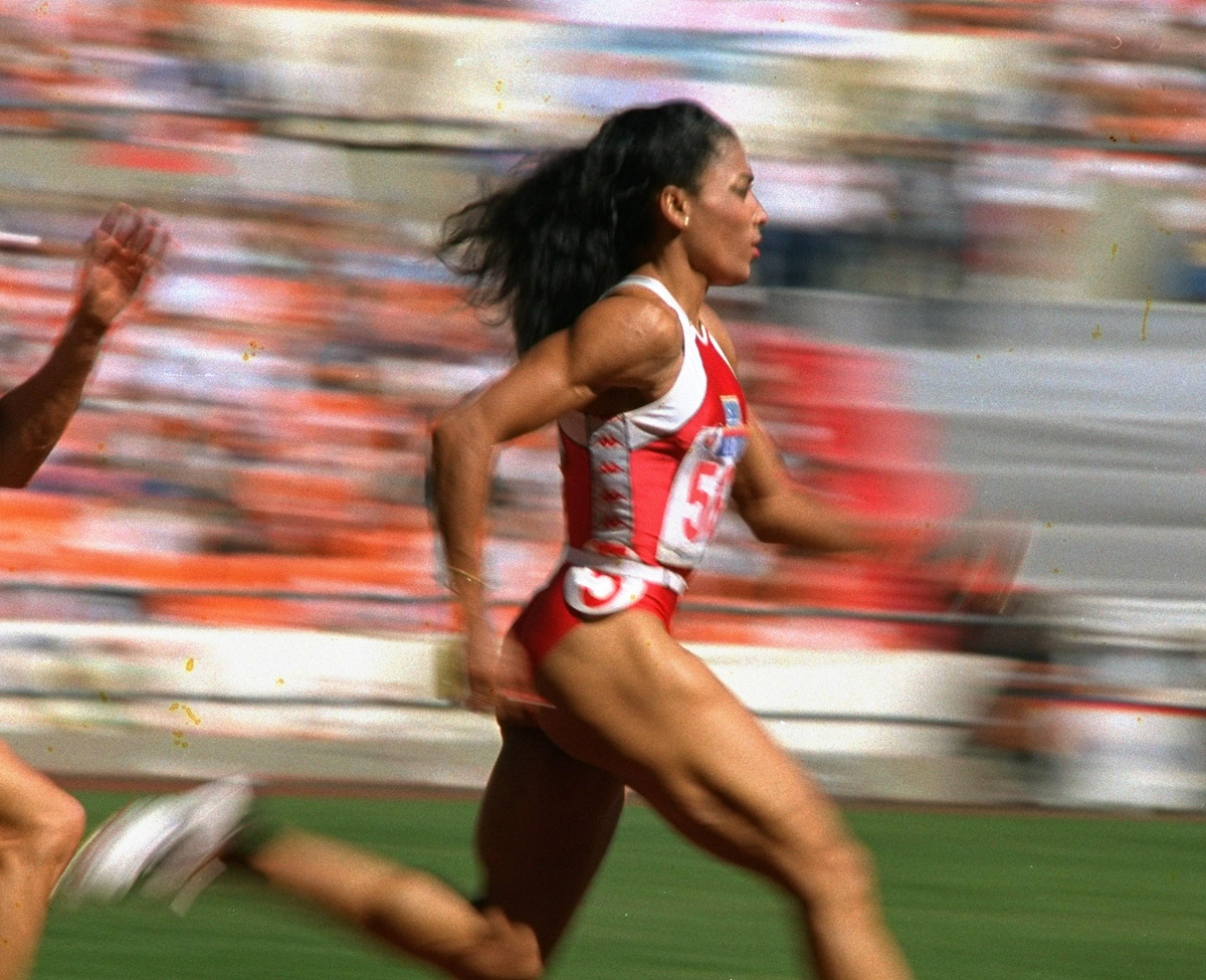 A female athlete in a red and white uniform sprints on a track during a race, with motion blur emphasizing her speed. Stadium seats and other runners are visible in the background. 9. Florence Griffith-Joyner’s 10.49 in the Women’s 100 Meters