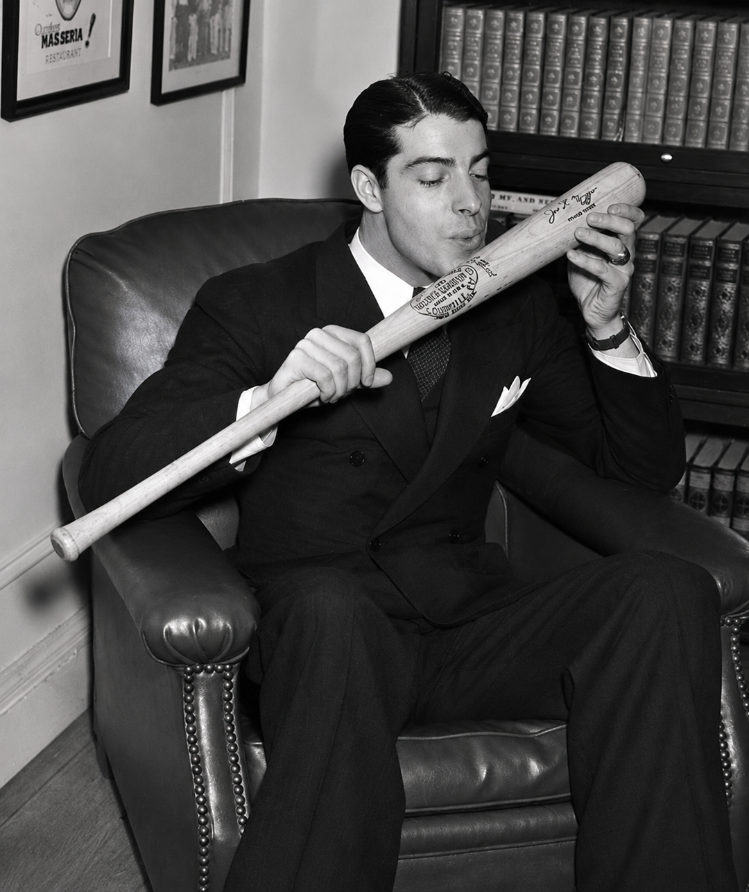 A man in a suit sits in an armchair and kisses a baseball bat. The bat has visible writing and signatures on it. Shelves filled with books and framed pictures are in the background. 4. Joe DiMaggio’s 56-Game Hitting Streak