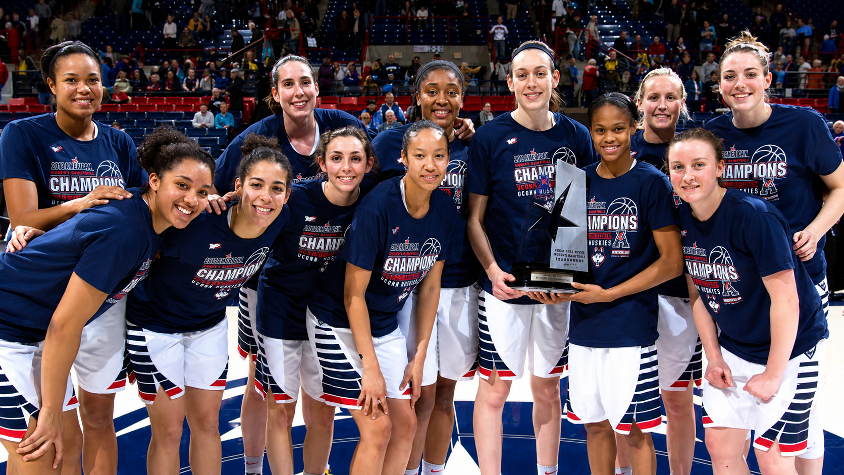 A women's basketball team poses together on a court, smiling and wearing matching "Champions" shirts. Several players hold a large trophy, celebrating a victory. Fans are visible in the background. 12. UConn Women’s Basketball’s 111-Game Winning Streak