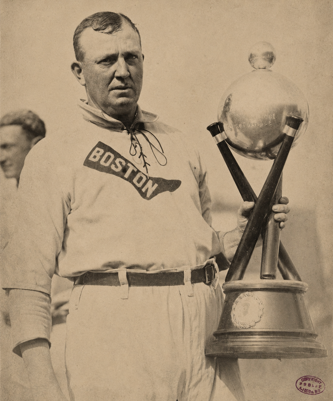 A vintage photo of a baseball player in a “BOSTON” jersey holding a large, ornate trophy with a globe on top. Another person is partially visible in the blurred background. 3. Cy Young’s 511 Major League Wins
