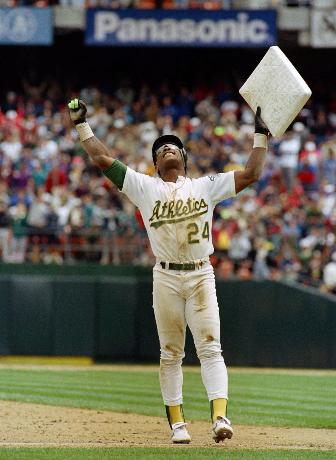 A baseball player in an Athletics uniform lifts a base triumphantly above his head on the field, celebrating as a crowd watches in the background. 11. Rickey Henderson’s 1,406 Stolen Bases