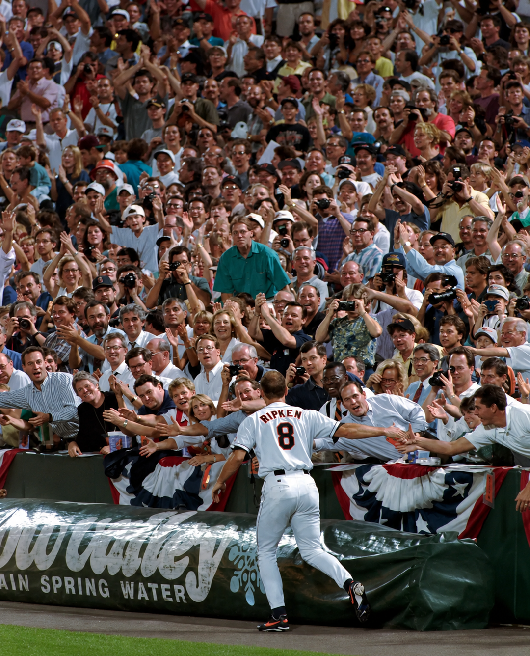 A baseball player wearing a number 8 “Ripken” jersey greets enthusiastic fans along the stands at a stadium, as a large crowd claps, waves, and takes photos. Patriotic bunting decorates the railing. 5. Cal Ripken Jr.’s 2,632 Consecutive Games