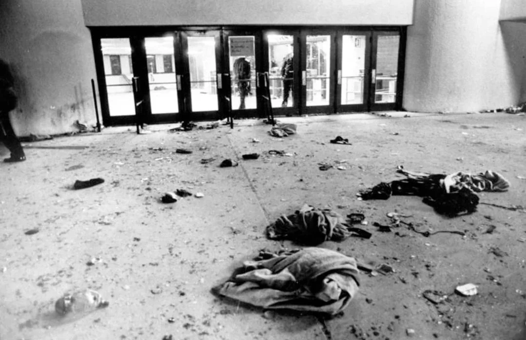 Black and white photo showing debris, clothing, and broken glass scattered outside a set of glass doors at a building entrance. The area appears deserted and damaged.