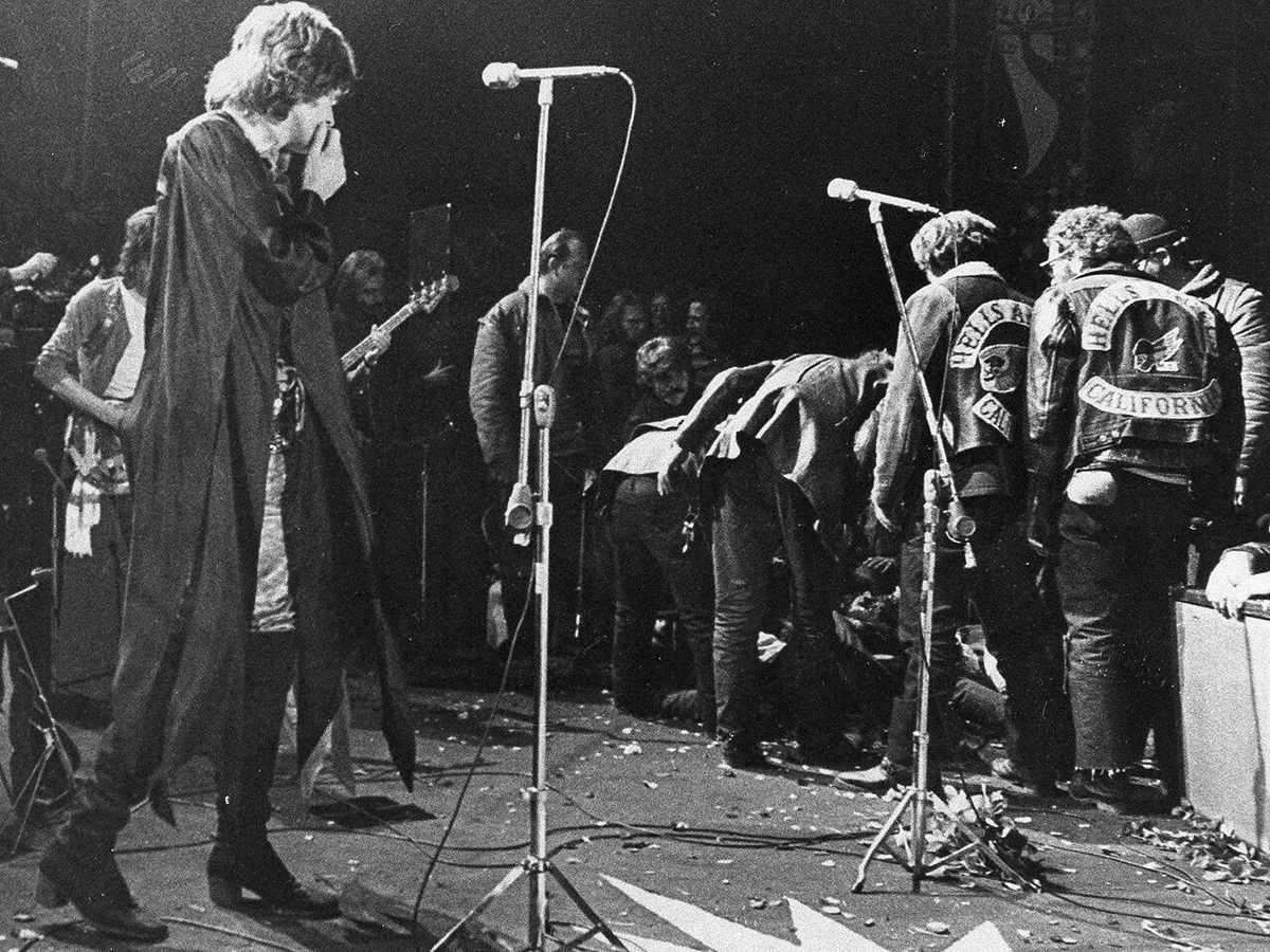 A black-and-white photo of a chaotic concert scene with a singer in a cloak covering his mouth and people, including men in "Hell's Angels" jackets, gathered around someone on the ground near microphones on stage.