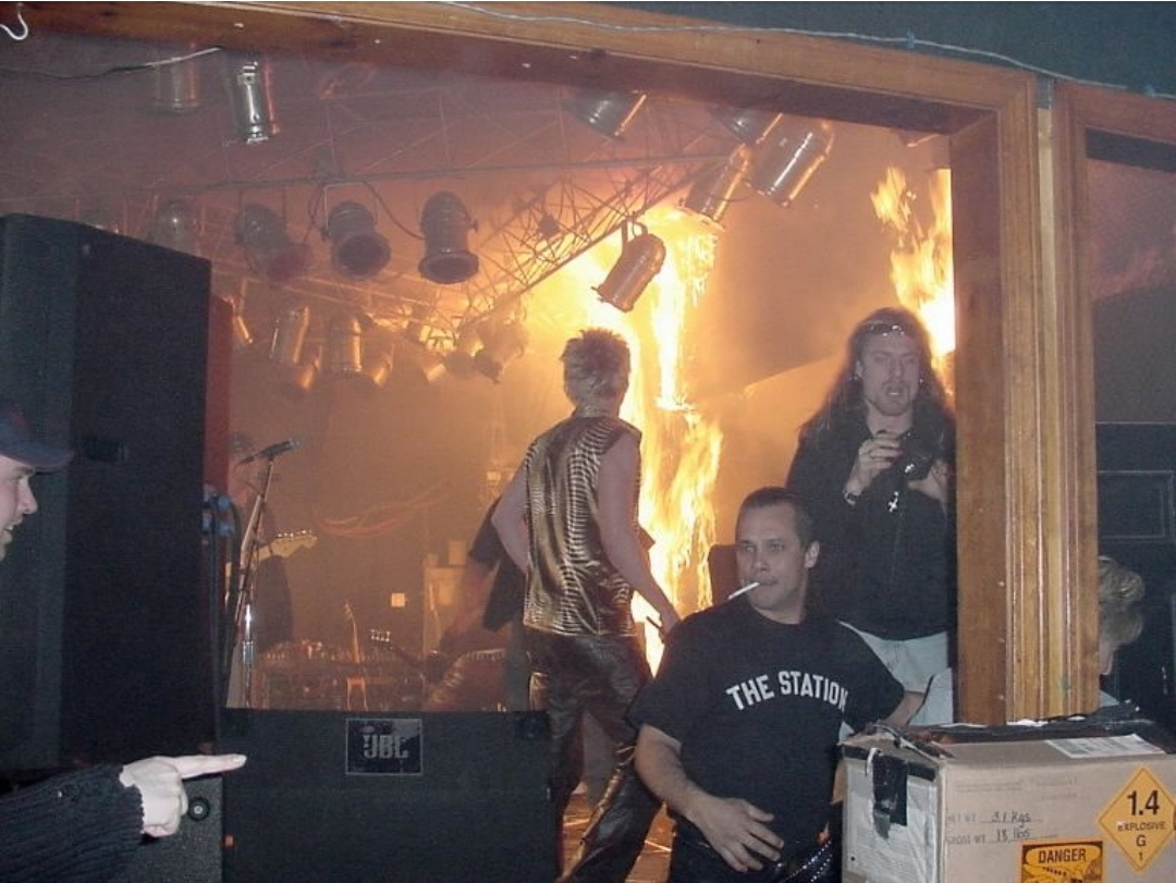 A band performs on stage as a fire blazes behind them, with flames reaching the ceiling. People appear alarmed, and one man wearing a shirt that reads "The Station" stands in the foreground near a box labeled "Danger.
