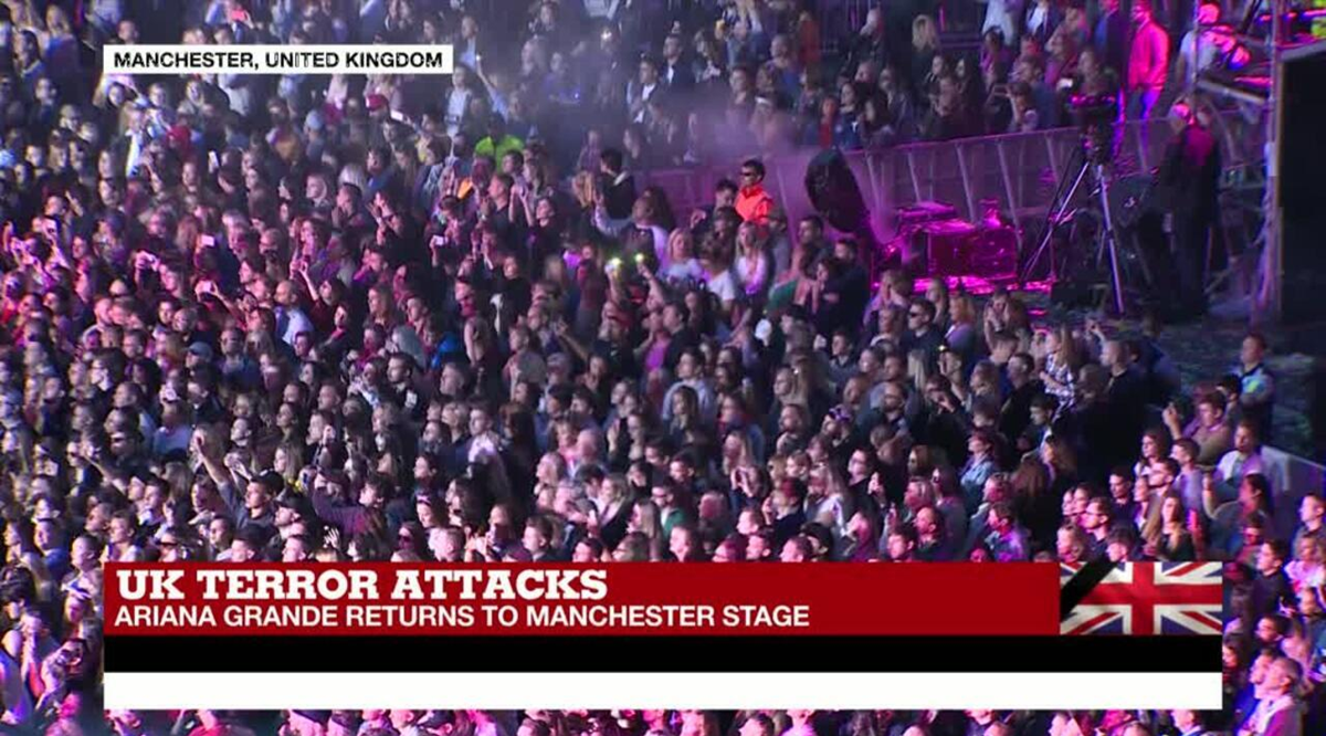 A large crowd attends a concert in Manchester, UK, at night. A news banner reads, "UK Terror Attacks—Ariana Grande returns to Manchester stage," with the UK flag in the corner.