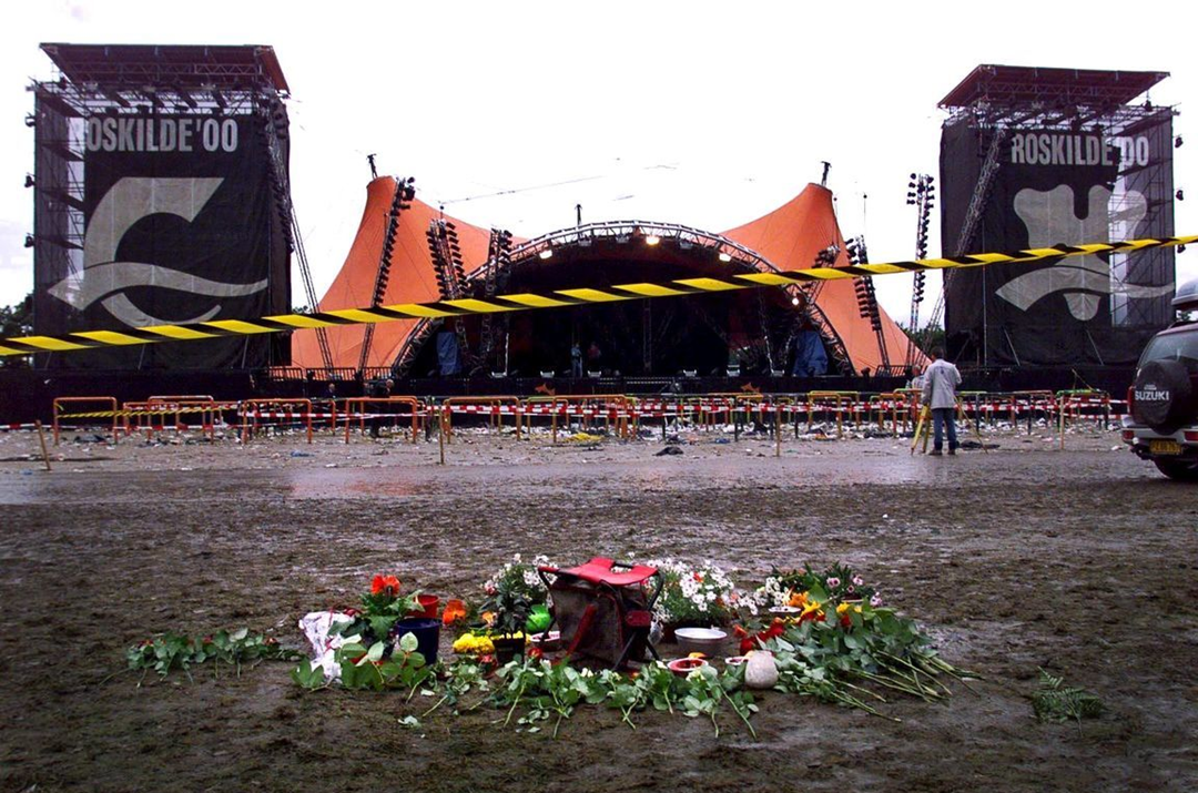 A muddy festival stage with "Roskilde '00" banners is seen in the background. In the foreground, a memorial of flowers, candles, and a backpack marks the ground, surrounded by caution tape. The sky is overcast.