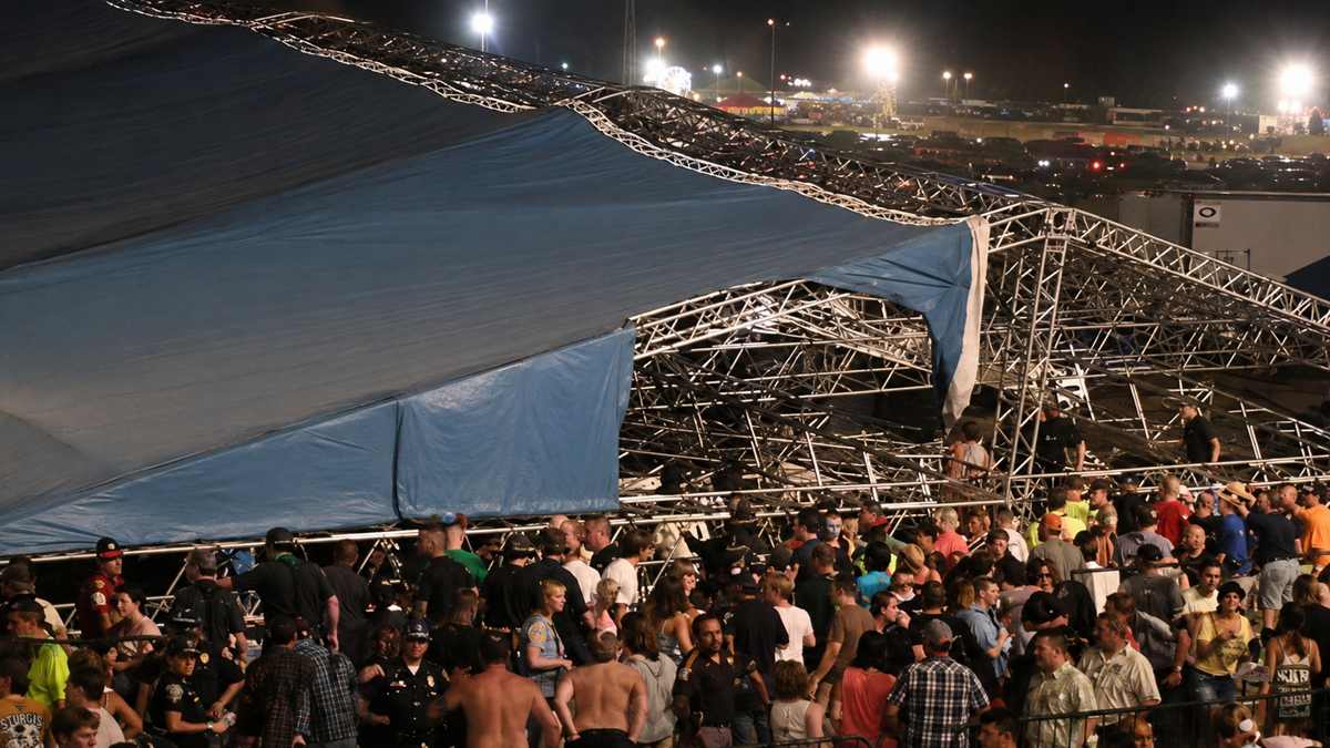 A large crowd gathers under a partially collapsed stage structure with twisted metal beams and a sagging blue tarp at night, while emergency personnel and spectators look on.