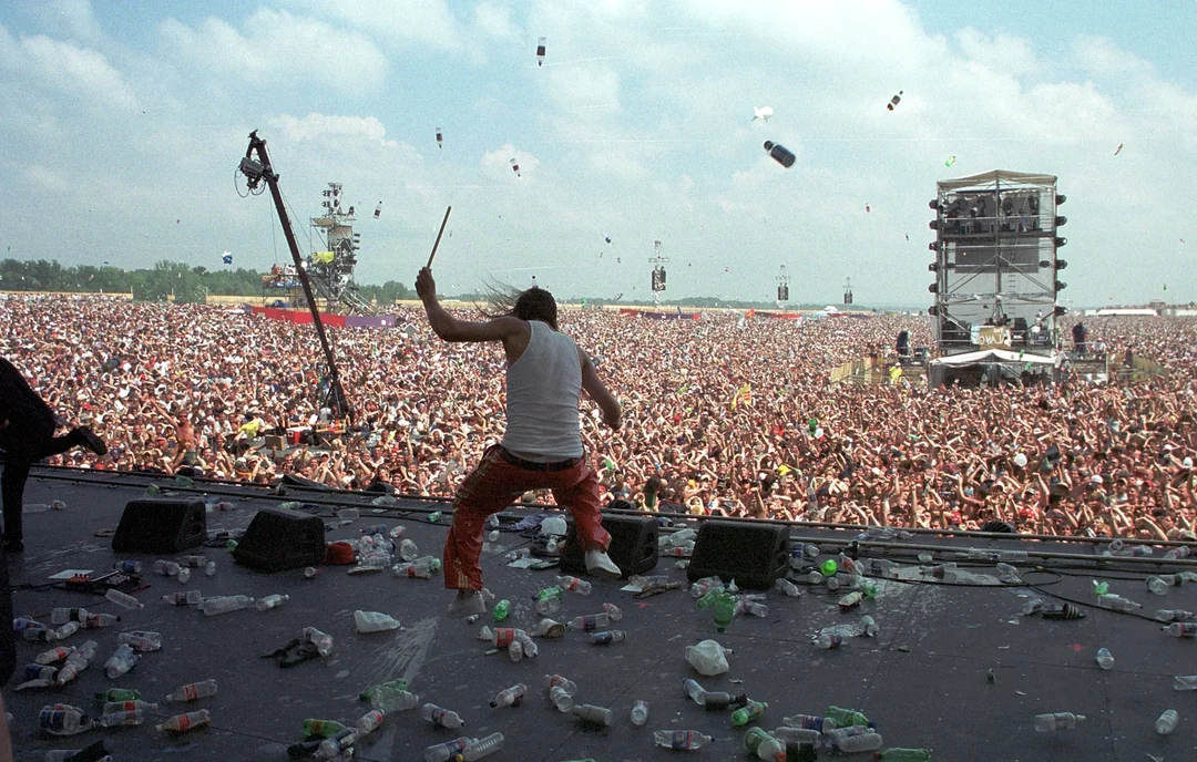 A musician on stage faces a massive crowd at an outdoor concert, with plastic bottles littering the stage and many flying through the air under a partly cloudy sky.