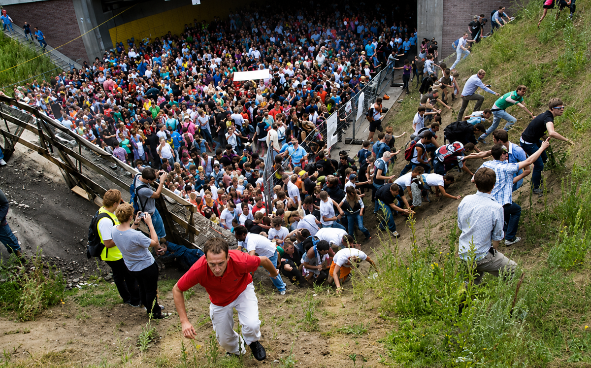 A large crowd of people gathers at the bottom of a grassy hill; many climb up the slope, some helping others, while more people watch or follow behind, creating a scene of urgency and movement.