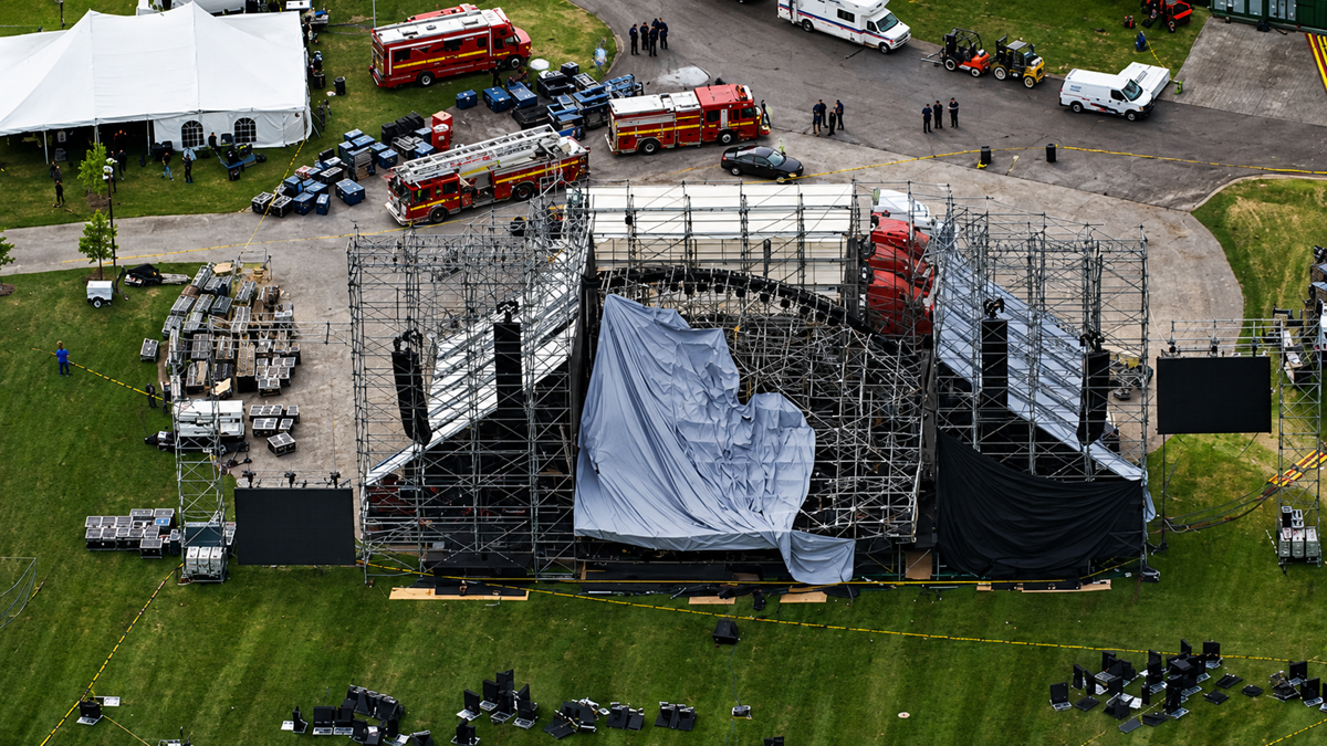 Aerial view of a collapsed outdoor concert stage with metal scaffolding, tarps, and equipment scattered on the grass. Emergency vehicles and personnel are present, and the area is cordoned off.