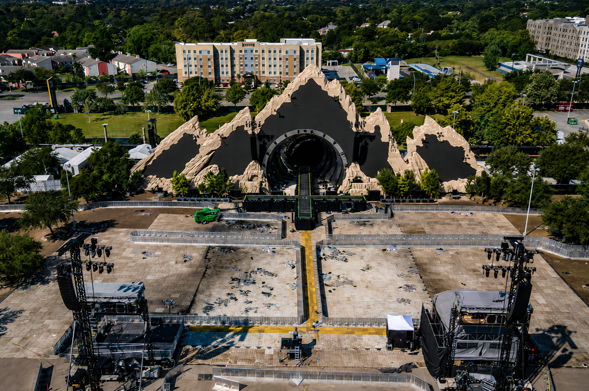 A large outdoor concert stage with a mountain-like backdrop and a central circular opening is set up in an empty lot, with lighting towers and equipment visible. Trees and buildings are in the background.