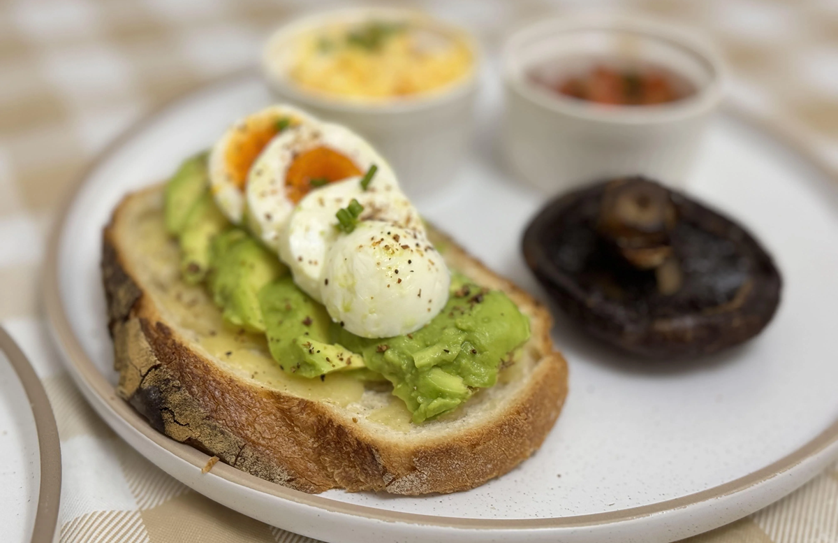 A plate with avocado toast topped with sliced boiled eggs and herbs, accompanied by a grilled mushroom and two small bowls containing scrambled eggs and salsa.