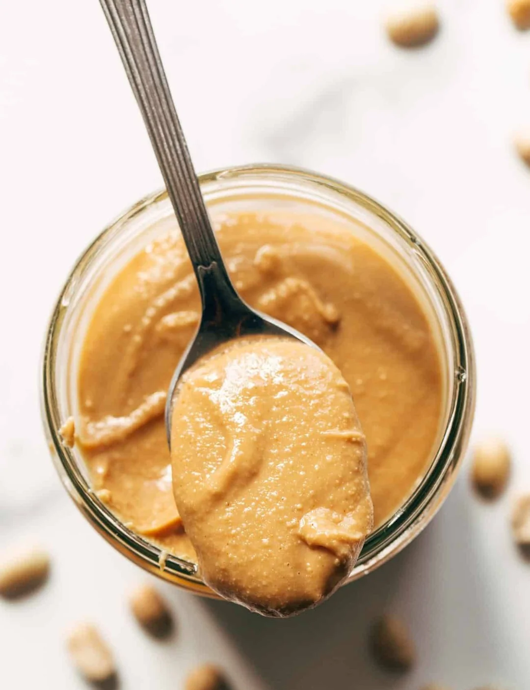 A close-up view of a jar filled with creamy peanut butter, with a metal spoon scooping out a portion. Whole peanuts are scattered on the white surface around the jar.