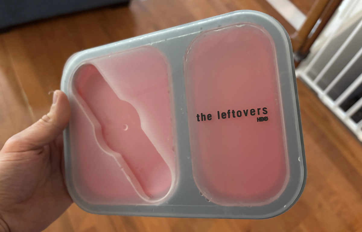 A hand holds a pink and gray frozen popsicle mold with "the leftovers HBO" printed on it. The mold is divided into two sections and rests over a hardwood floor.
