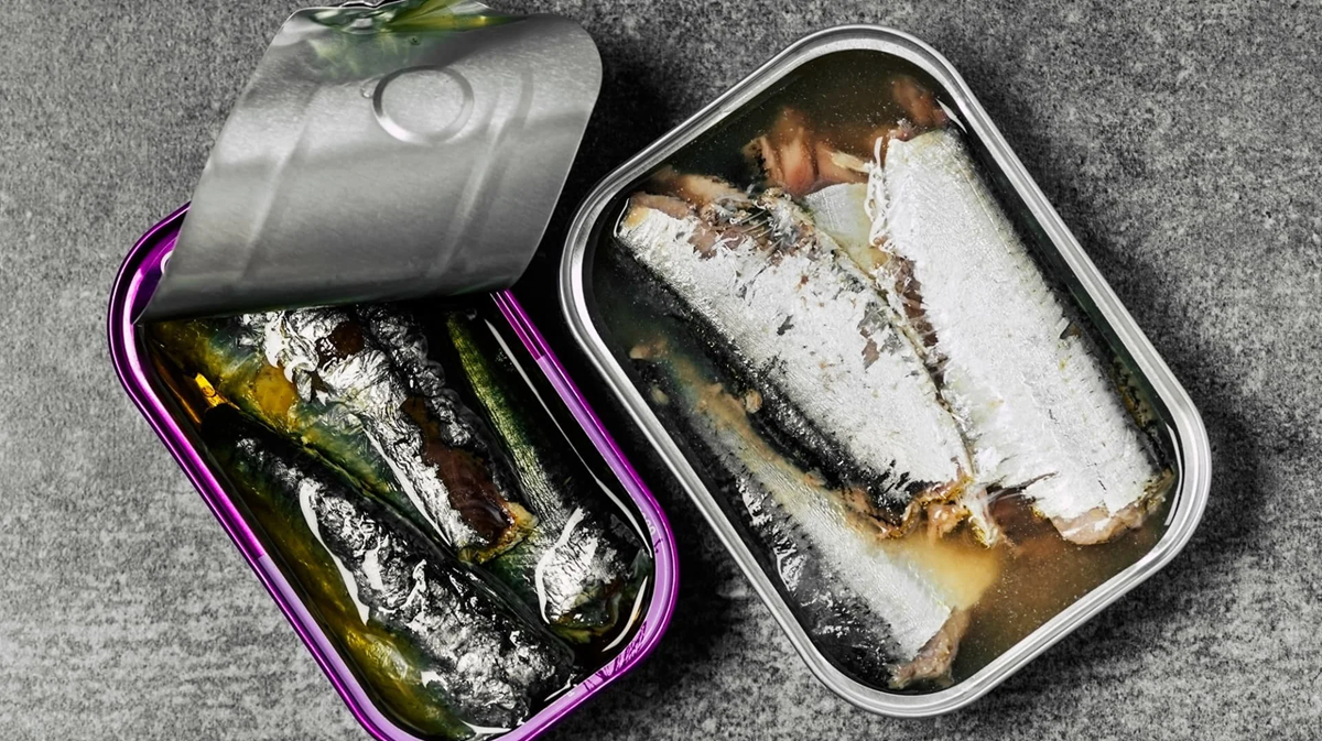 An open tin can filled with sardines in oil sits on a gray surface, with the lid partially peeled back to reveal several shiny, silvery fish packed closely together.